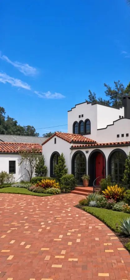 AI concept rendering of a curving red brick paver drive lined with tropical plantings leading to a white Spanish Revival home