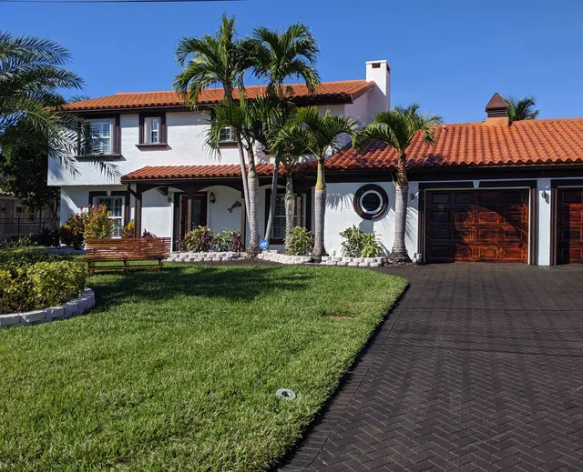 White two-story home with a red tile roof, dark wood garage doors, a round porthole window, palms and a herringbone paver drive