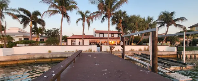 View from a wooden dock at sunset toward a white stucco home with a red tile roof behind a seawall