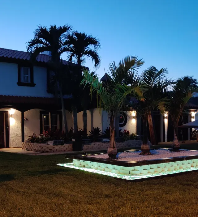Raised palm planter with glowing block edging in front of a white home at dusk