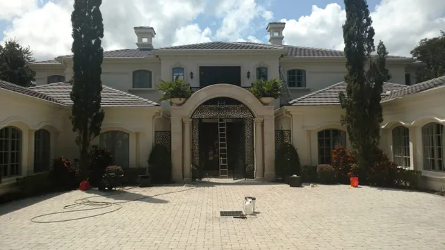 Cream stone estate with an arched iron entry, a ladder at the door and pressure-washing hoses and equipment on the paver drive