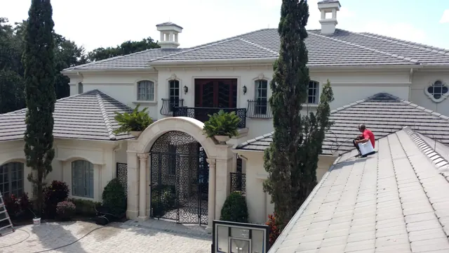 Wider view of a cream stone estate with a technician in a red shirt on the tile roof and a ladder at the side of the house