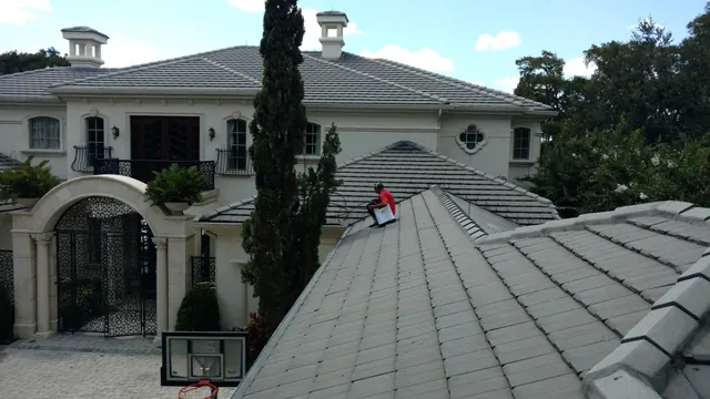 Technician in a red shirt working on a flat tile roof of a cream stone estate with an arched iron entry below