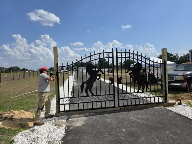 Installer in a red cap fitting a black iron double gate with rearing-horse silhouettes at the end of a newly paved drive with white rock borders