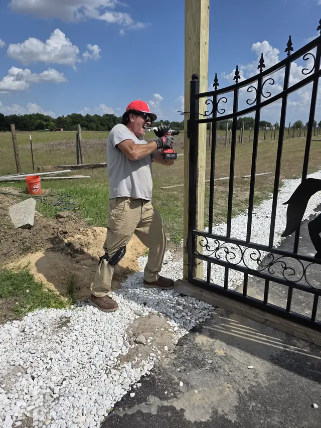 Worker in a red cap using a drill while installing a black ornamental iron gate on a wood post