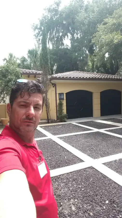 Man in a red shirt taking a selfie on a paver-grid drive in front of a yellow garage with arched black doors