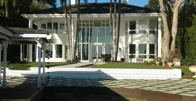 White lakefront home with a diamond-pattern paver terrace, white retaining wall steps and palms