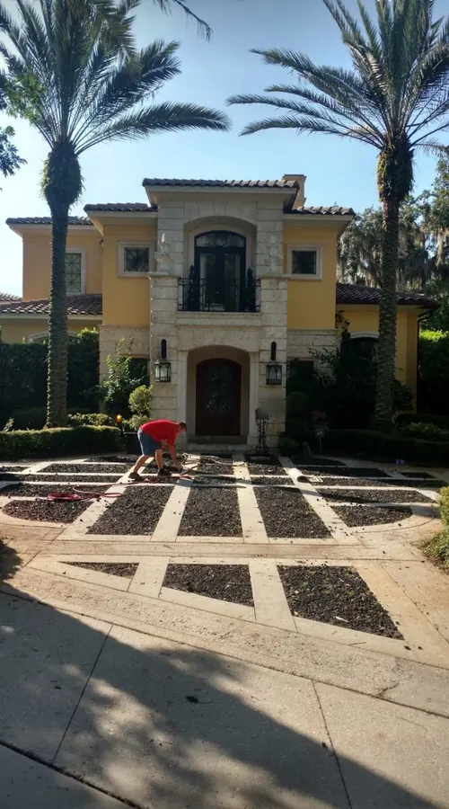 Crew member in a red shirt kneeling on a paver-grid drive of stone bands and dark gravel in front of a yellow Mediterranean estate